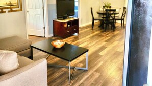 A living room with a sofa, a coffee table with a bowl, a TV on a stand, a dining area with chairs and a table, and a refrigerator. - Greenhill Boutique Suites (Newport News)
