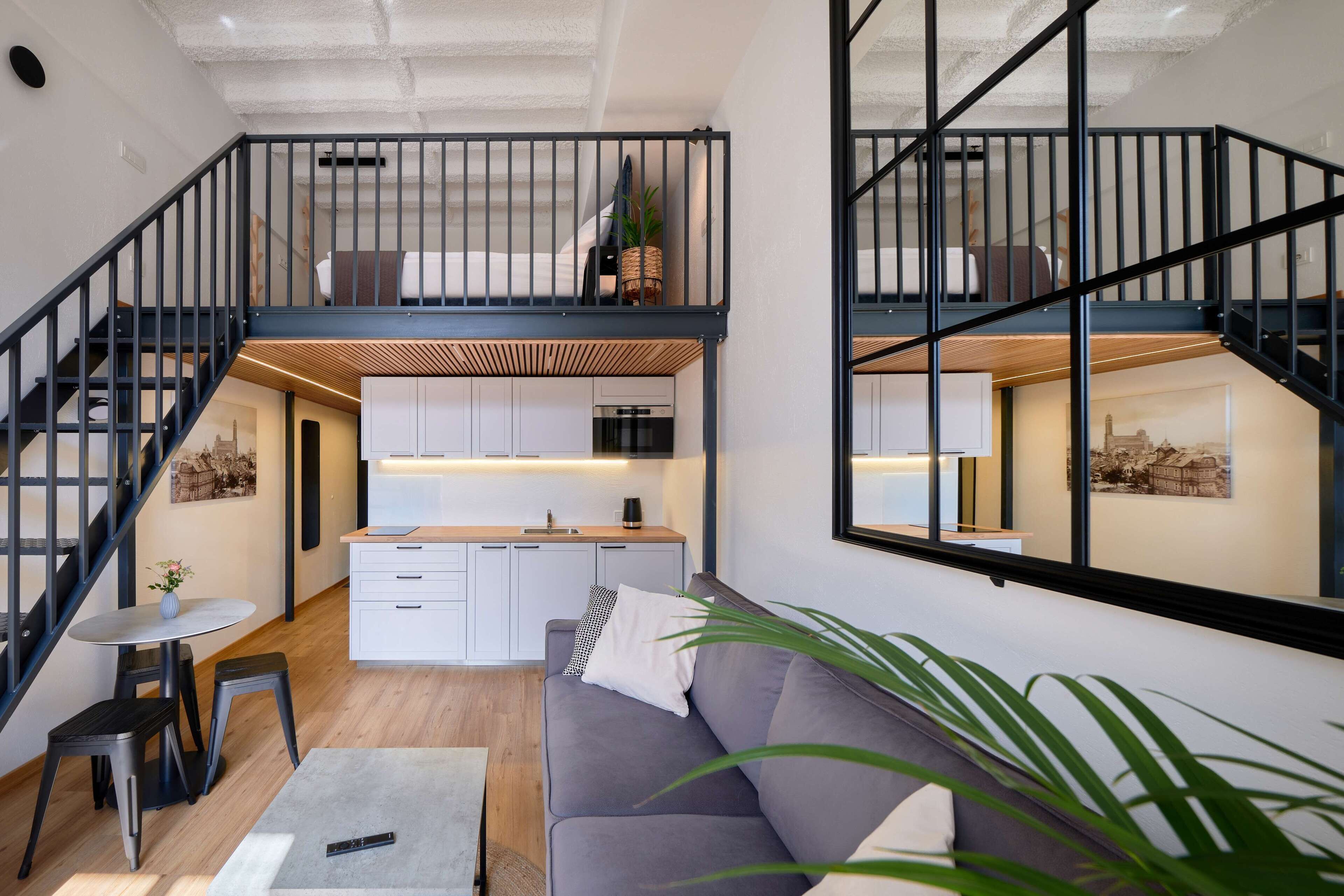 Un loft moderno con una escalera que sube a una zona de cocina elevada, un sofá gris y una pequeña mesa de comedor.