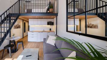 Un loft moderno con una escalera que sube a una zona de cocina elevada, un sofá gris y una pequeña mesa de comedor.