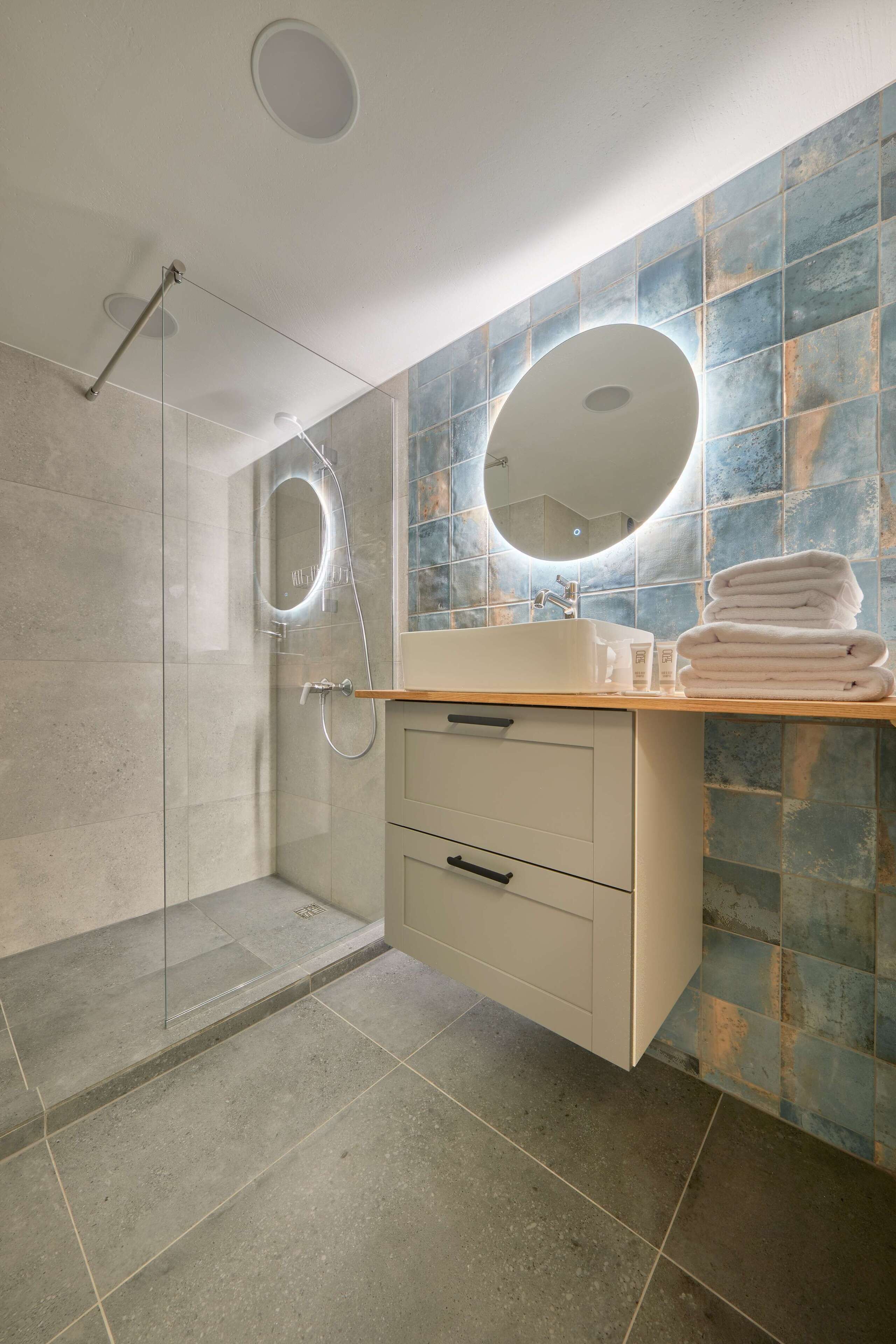 Un baño moderno con ducha envuelta en vidrio, un lavabo con espejo redondo y pared de azulejos.