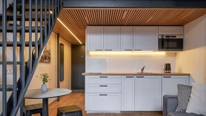 A modern kitchen with white cabinets, a built-in oven, and a wooden ceiling. There is a round table with stools and a sofa with a patterned pillow. - Queen Barbora Central Lofts (Kaunas)