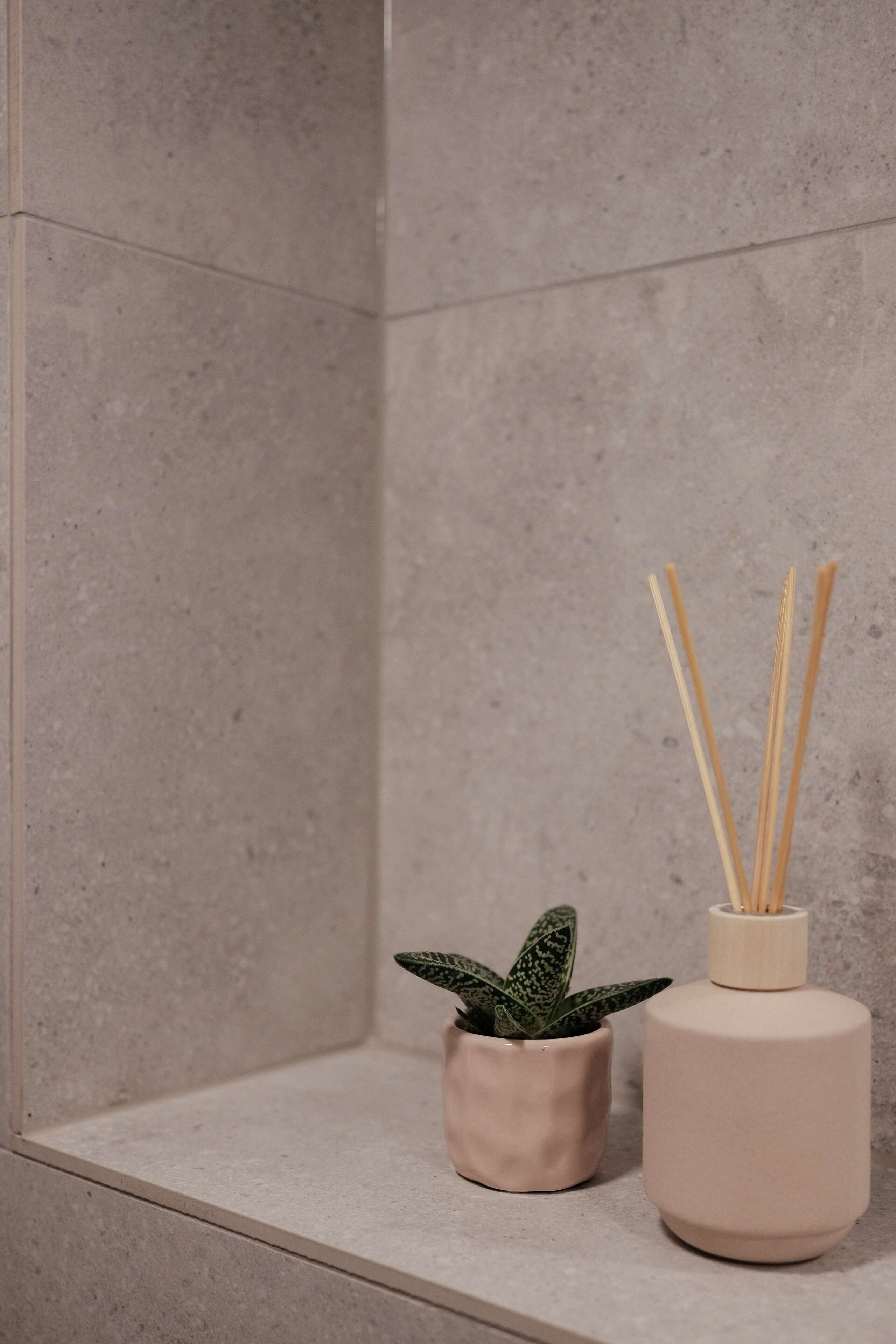 Casa de banho com parede revestida a azulejos bege, uma pequena planta em vaso e um recipiente cilíndrico com raminhos de cana.