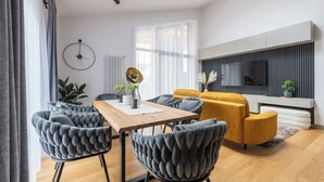 A modern living room with a wooden dining table, tufted grey chairs, a mustard yellow sofa, and a flat-screen TV mounted on the wall. - Luxurious penthouse with parking
 (Riga)