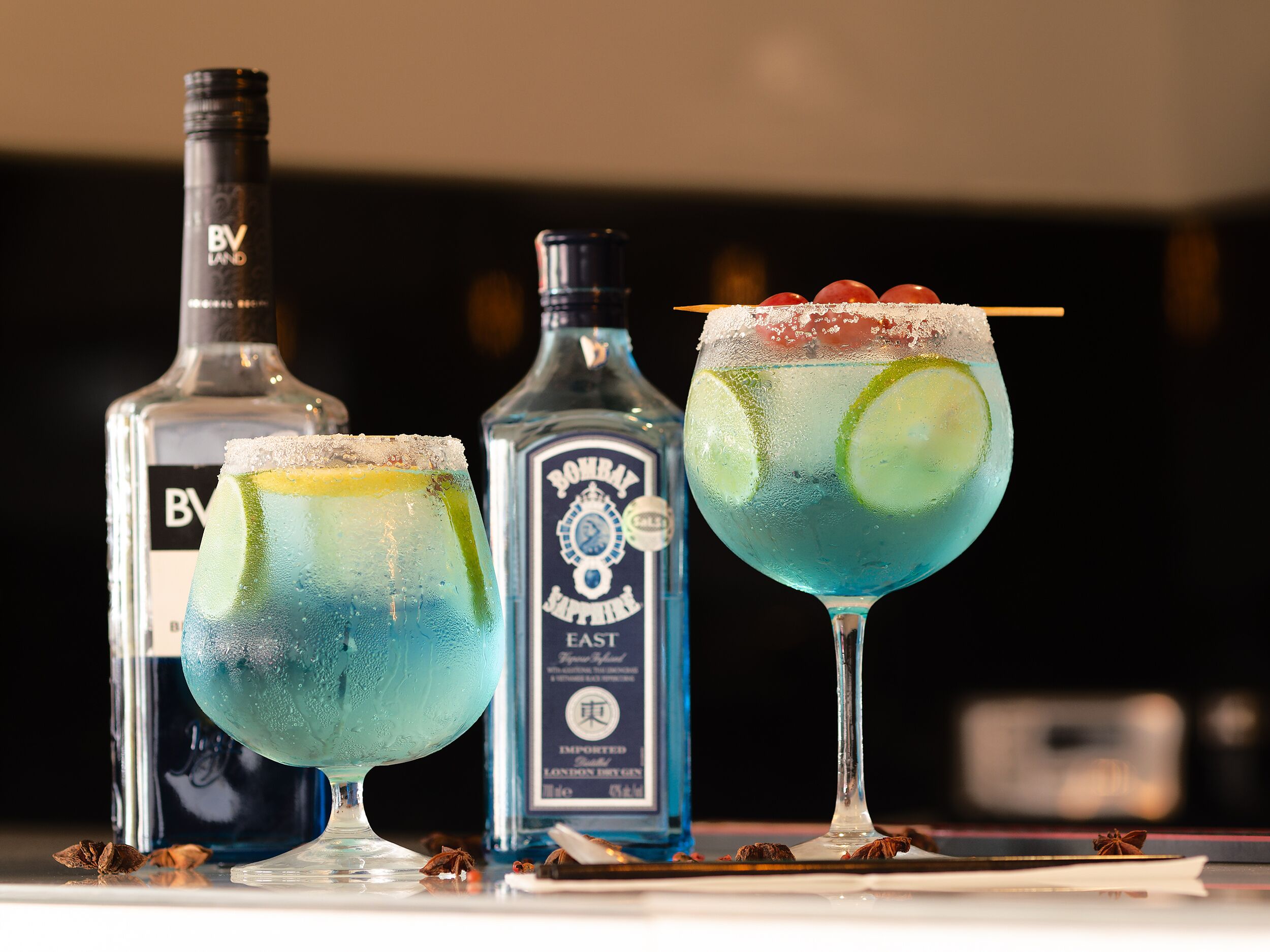 Drie cocktails met garnering, een fles Bombay Sapphire gin en een fles B.V. Land wodka.