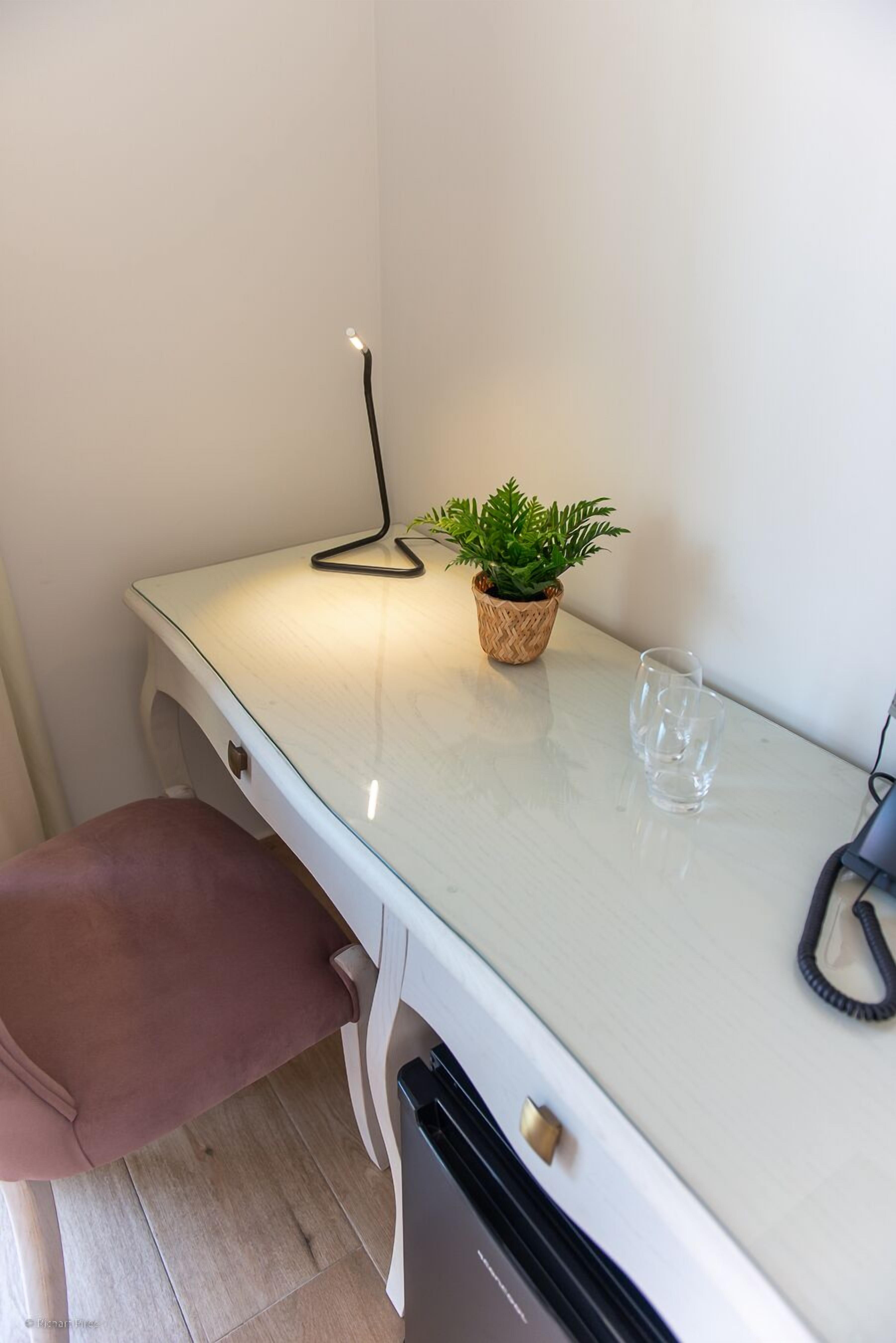 A small, white desk with a plant, a glass, and a lamp.
