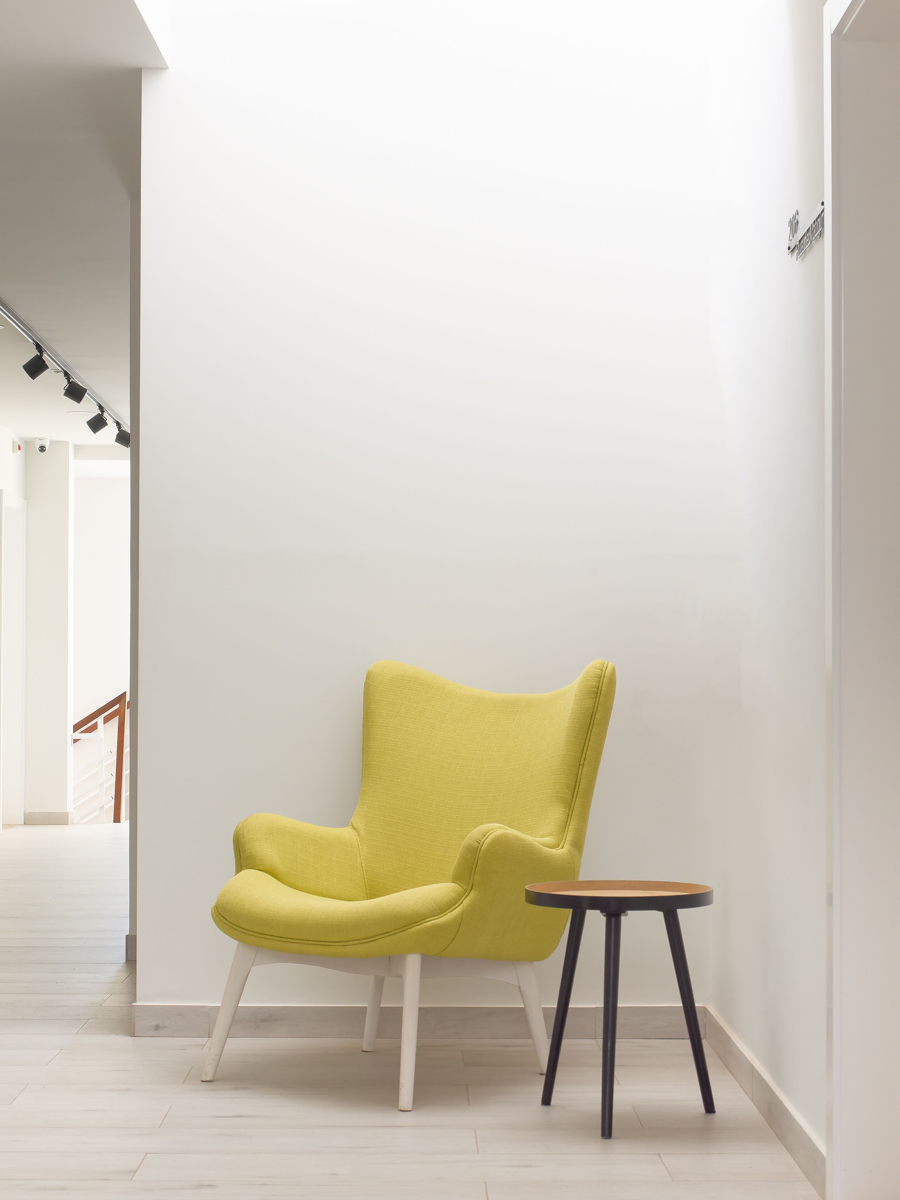 A bright yellow armchair with white legs, positioned next to a small round table with a wooden top and black legs, in a hallway with white walls and a wooden floor.