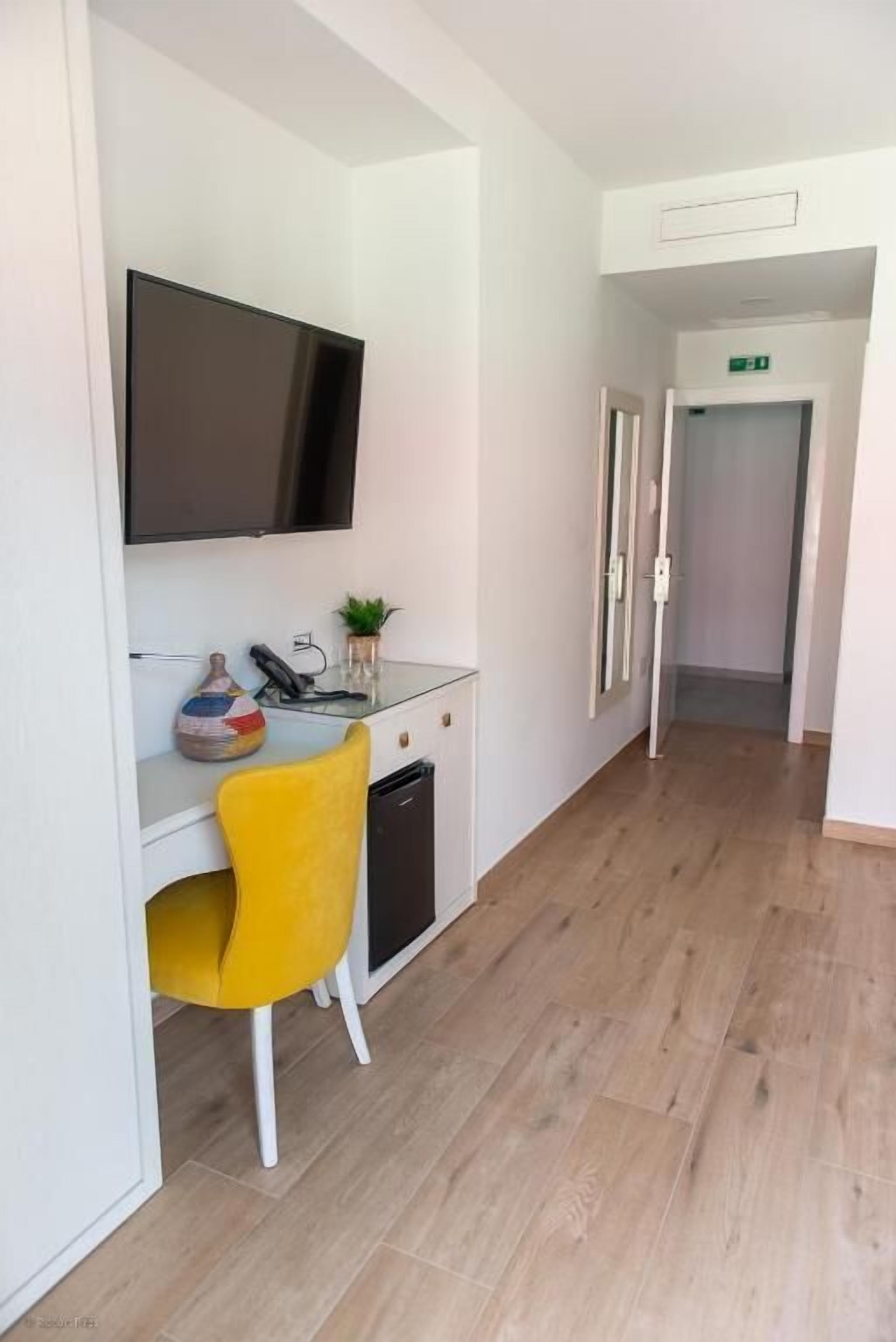 Una habitación de hotel moderna con suelo de madera, una silla amarilla, un escritorio de cristal con televisión de pantalla plana, una planta pequeña y un florero.
