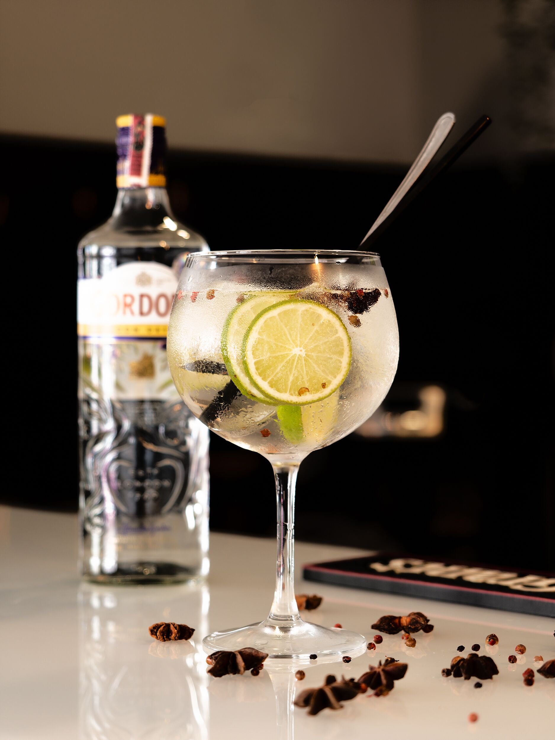 A cocktail glass with lime slices and spices, a bottle of liquor, and a spoon on a reflective surface.