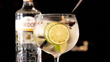A cocktail glass with lime slices and spices, a bottle of liquor, and a spoon on a reflective surface.