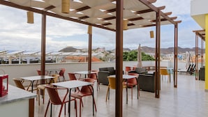 Una terraza al aire libre con mesas y sillas, una pérgola y vistas a edificios y montañas.