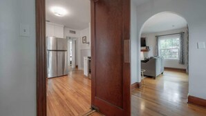 A modern kitchen with stainless steel appliances, a wooden floor, and an open layout leading to a living area with a grey sofa and a window.