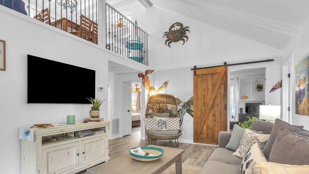 A spacious living room with a high ceiling, a large flat-screen TV mounted on the wall, a wooden dining table, a comfortable sofa, and a decorative chair. - Tipsy Turtle: Warm and Inviting, Short Walk to the Beach (Oak Island)