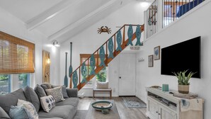 A living room with a grey sofa, a coffee table, a staircase with a fish railing, and a television mounted on the wall. - Tipsy Turtle: Warm and Inviting, Short Walk to the Beach (Oak Island)