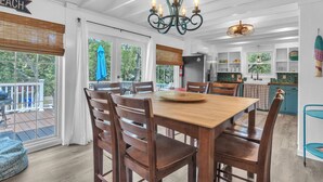 A spacious dining area with a wooden table and chairs, a chandelier, and large windows offering a view of the outdoors. - Tipsy Turtle: Warm and Inviting, Short Walk to the Beach (Oak Island)