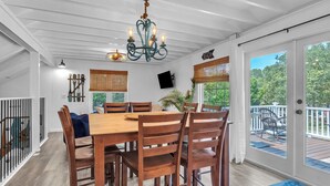 A dining area with a wooden table and chairs, a chandelier, and a view of the outdoors. - Tipsy Turtle: Warm and Inviting, Short Walk to the Beach (Oak Island)
