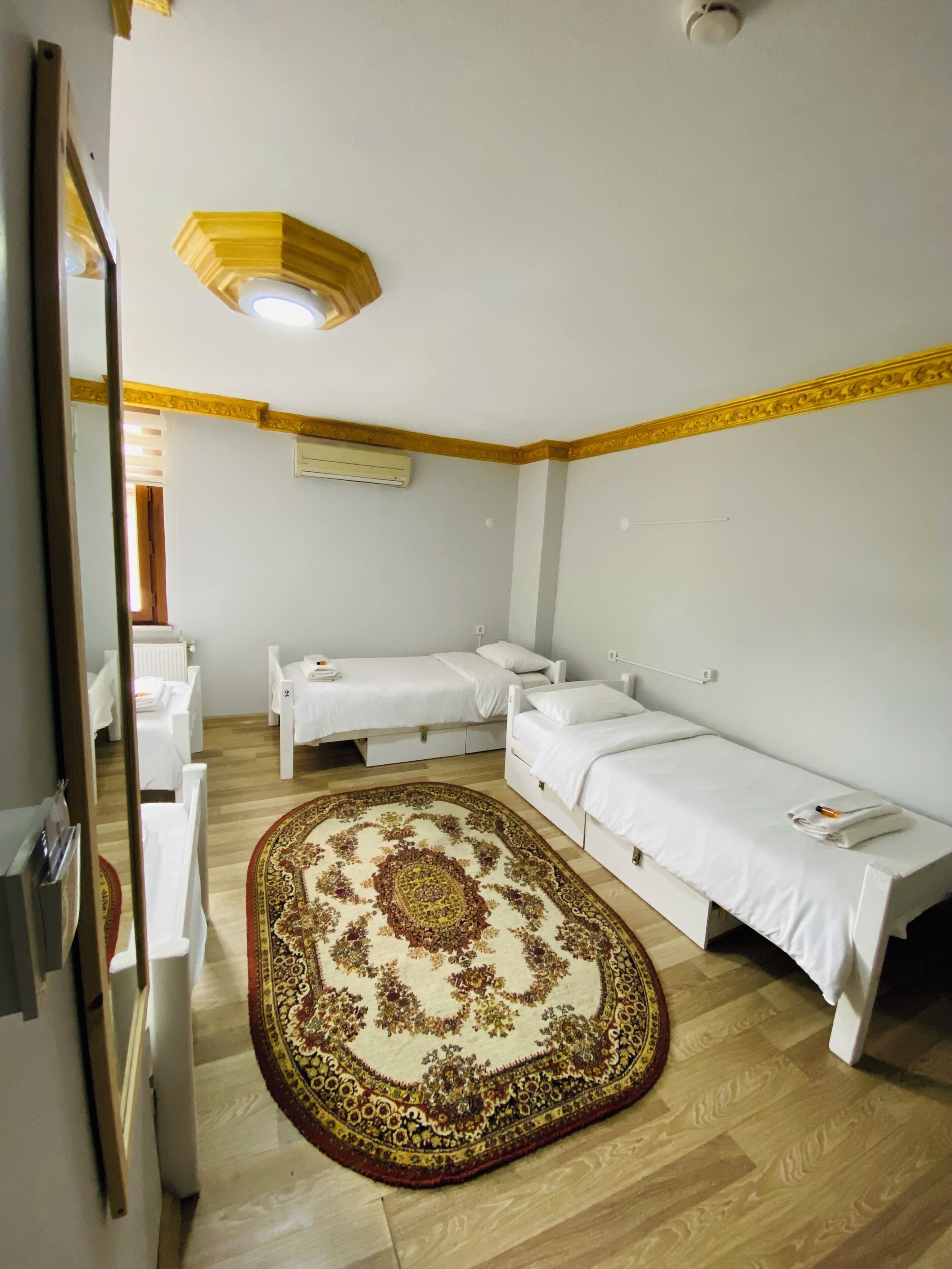 Ett hotellrum med två sängar, en stor, utsmyckad matta, en taklampa och en vit luftkonditioneringsenhet.