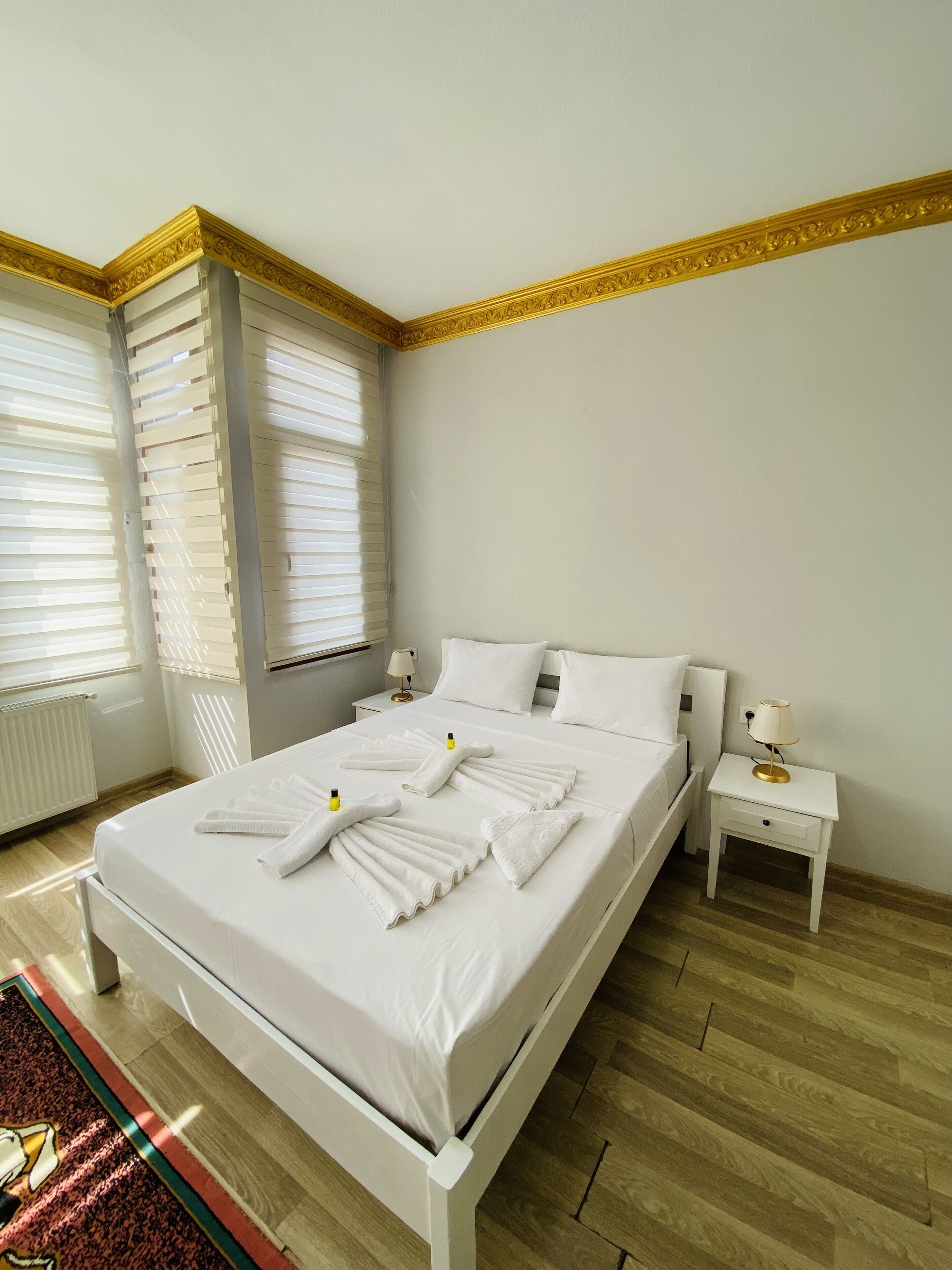 A neatly made bed with white linens and towels, a bedside table with a lamp, and a window with blinds.