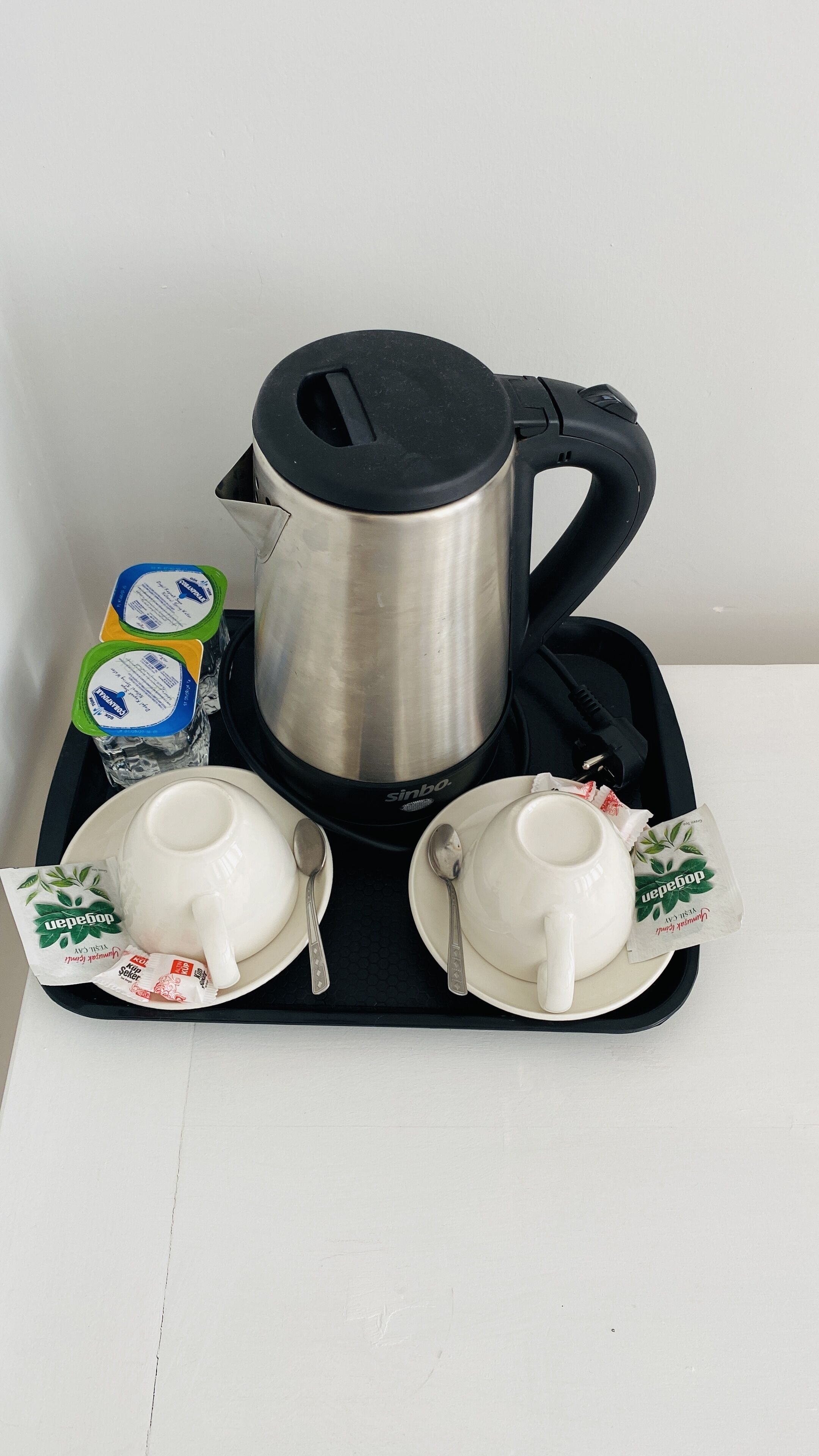 Een roestvrijstalen koffiezetapparaat met een zwarte handgreep, twee witte kopjes, twee lepels en twee yoghurtbekers op een dienblad.