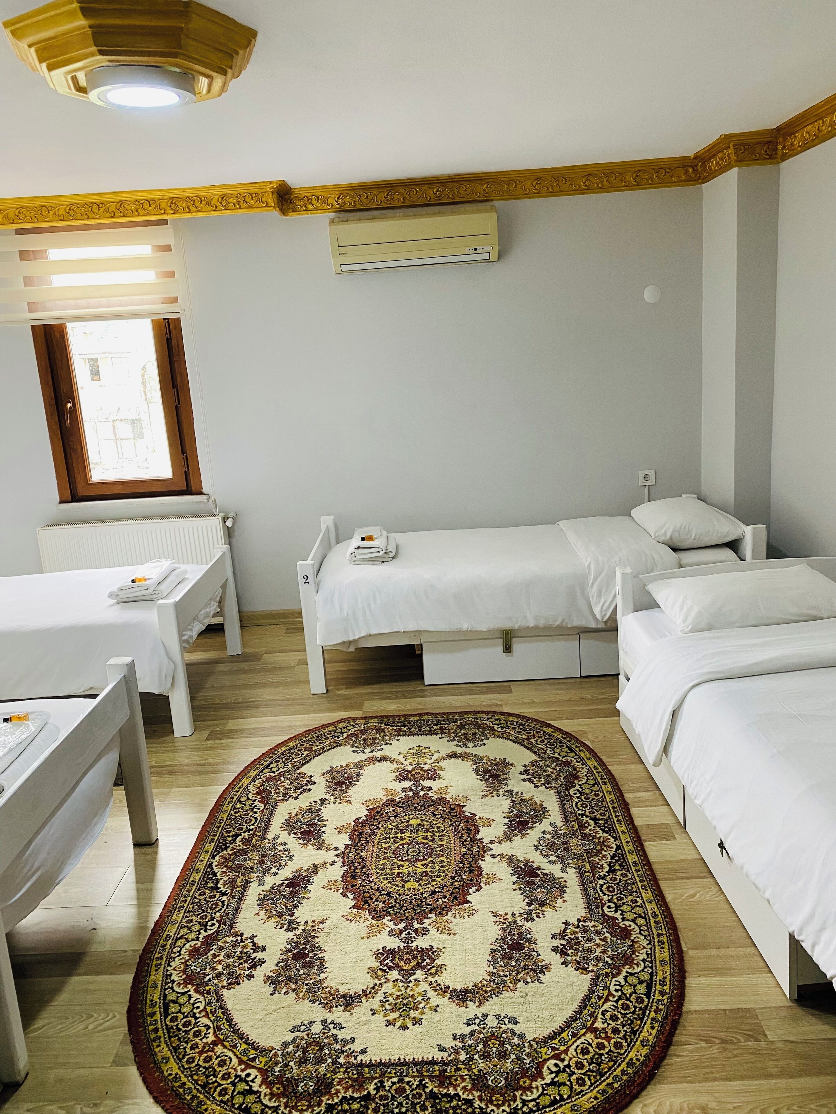 A hotel room with two single beds, a large patterned rug, a ceiling light, and a window with blinds.