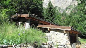 A rustic stone building with a sloped roof, surrounded by lush greenery and a mountainous backdrop. - Chalet de Bébert (Conthey)