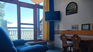 Una habitación con un ventanal amplio, un sofá, una mesa de madera con sillas y un televisor montado.