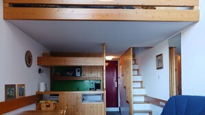 Habitación compacta con una mesa de comedor de madera, una silla azul, kitchenette con microondas y una escalera que conduce a otra zona.
