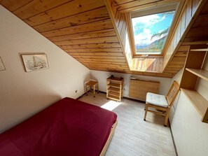 Ein Schlafzimmer mit schrägen Wänden, einem Oberlicht, einem Bett, einem Sessel, einem kleinen Tisch und einem Holzschrank.