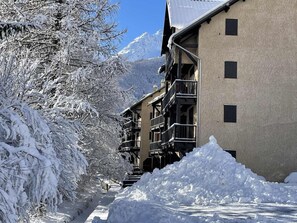 Ein schneebedecktes Gebäude mit mehreren Balkonen, ein verschneiter Weg und ein Berg im Hintergrund.