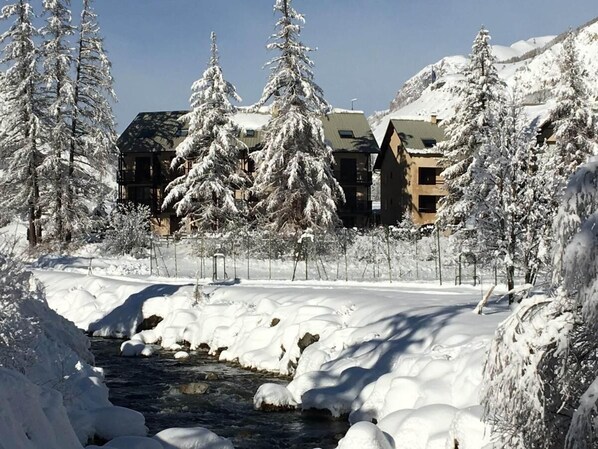 Eine verschneite Landschaft mit Fluss, Gebäuden und schneebedeckten Bäumen.