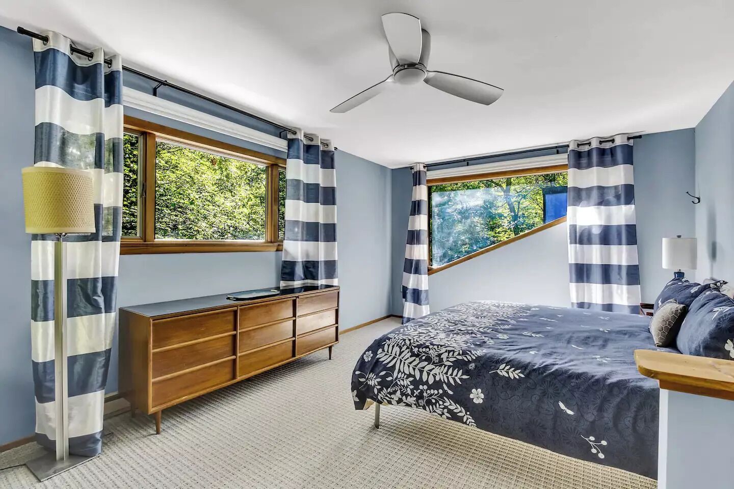 Une chambre à coucher avec un lit, une commode en bois, un ventilateur de plafond et une fenêtre avec des rideaux.