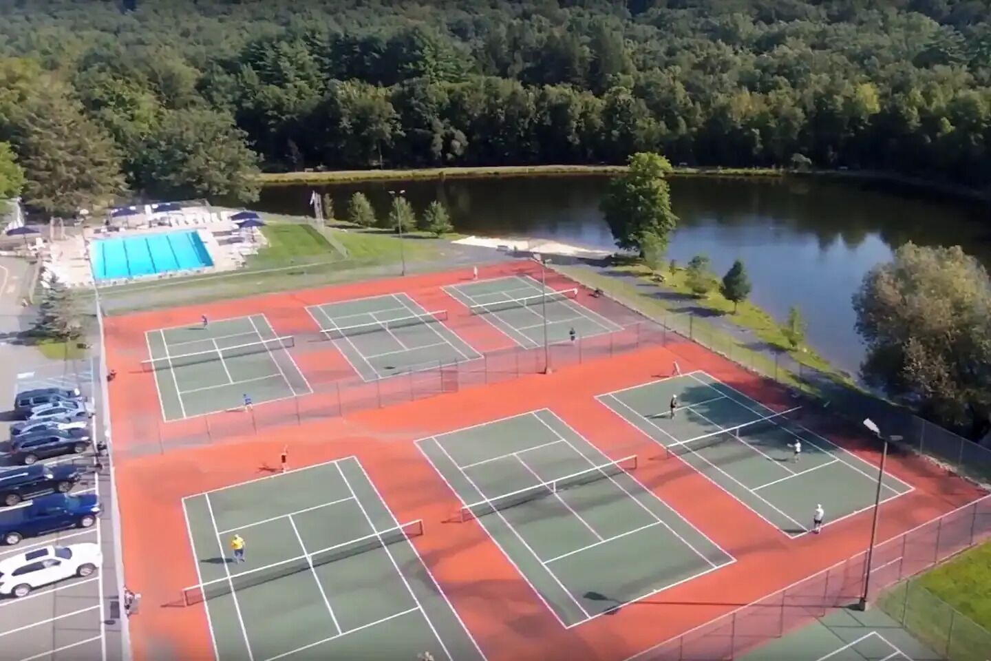 Un complejo de canchas de tenis con varias canchas, un estacionamiento y un lago al fondo.