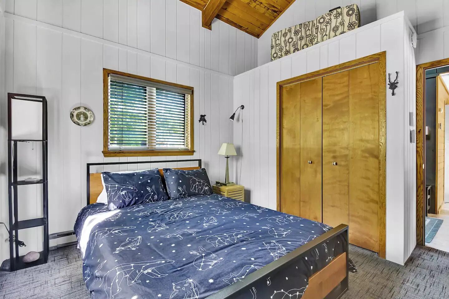 A bedroom with a bed, a window with blinds, a wardrobe, a bedside table with a lamp, and a wall-mounted decoration.