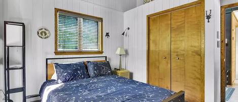 Un dormitorio con cama, ventana con persianas, armario, mesita de noche con lámpara y un adorno en la pared.