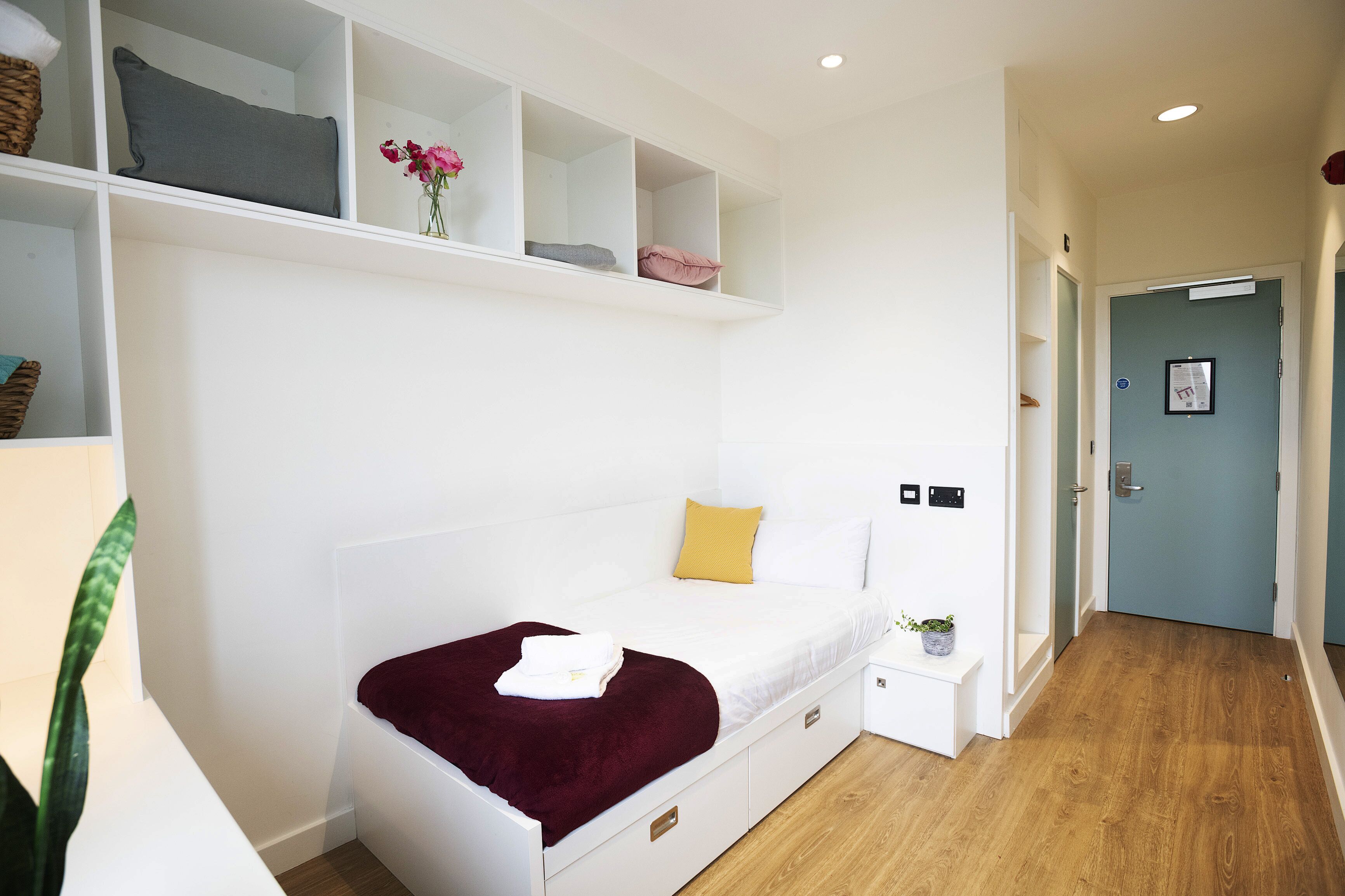 A bedroom with a bed, a bedside table, a shelf with pillows, and a plant.