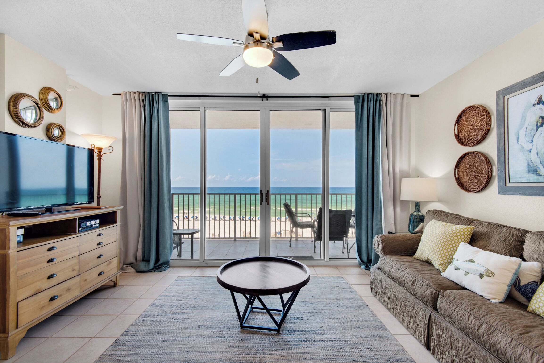 A living room with a television, a ceiling fan, a round coffee table, a sofa with cushions, and a view of the beach through the sliding glass door.
