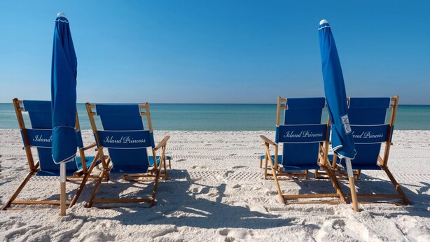 Four blue beach chairs with umbrellas on a sandy beach.