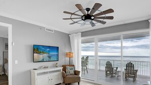 Habitación con ventilador de techo, televisor grande, una silla y balcón con vista al océano.