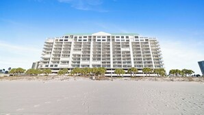 Un hotel de varios pisos con techo plano, situado en una playa arenosa con palmeras al frente.
