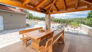Un patio al aire libre con una mesa de madera y bancos, una piscina al fondo y vistas del paisaje circundante.