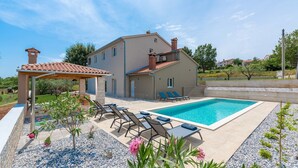 Una casa de dos pisos con piscina, mobiliario exterior y una pérgola.
