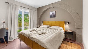 Un dormitorio con cama, mesita de noche, lámpara y vista a la vegetación por la ventana.