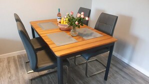 Een houten eettafel met vier stoelen, een vaas met bloemen, een schaal fruit en een waterfles.