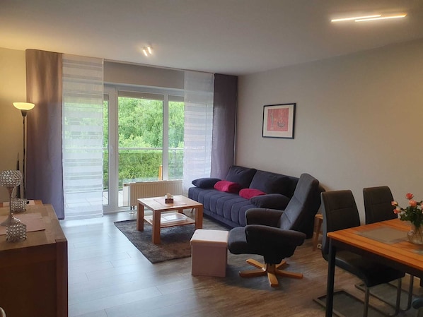 A modern living room with a sofa, a coffee table, a desk, and a chair. There is a large window with a view of greenery outside. - The most beautiful seagull shore on the North Sea coast, only 3 minutes walk to the beach (Wurster Nordseeküste)