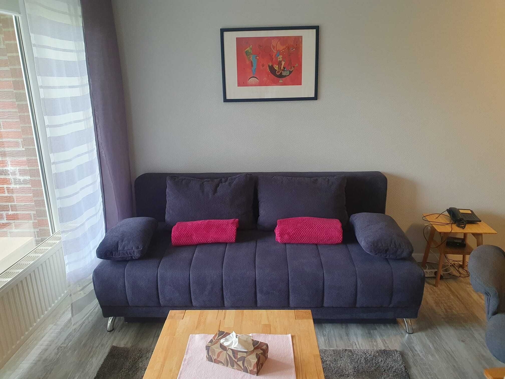 Un canapé bleu foncé agrémenté de deux coussins rouges, un tableau abstrait encadré au-dessus, et une petite table en bois sur laquelle se trouve un téléphone.