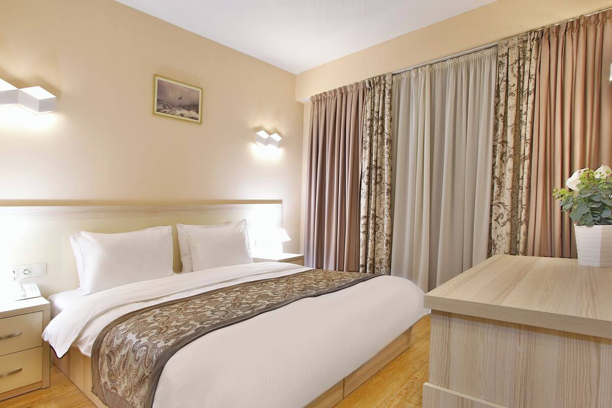 Habitación de hotel con cama, mesitas de noche, lámpara de pared, un cuadro y ventana con cortinas.