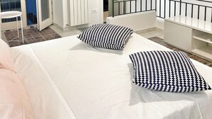 Une chambre à coucher avec un lit, deux coussins à motifs, un luminaire suspendu, une rambarde blanche et une porte donnant sur un balcon.