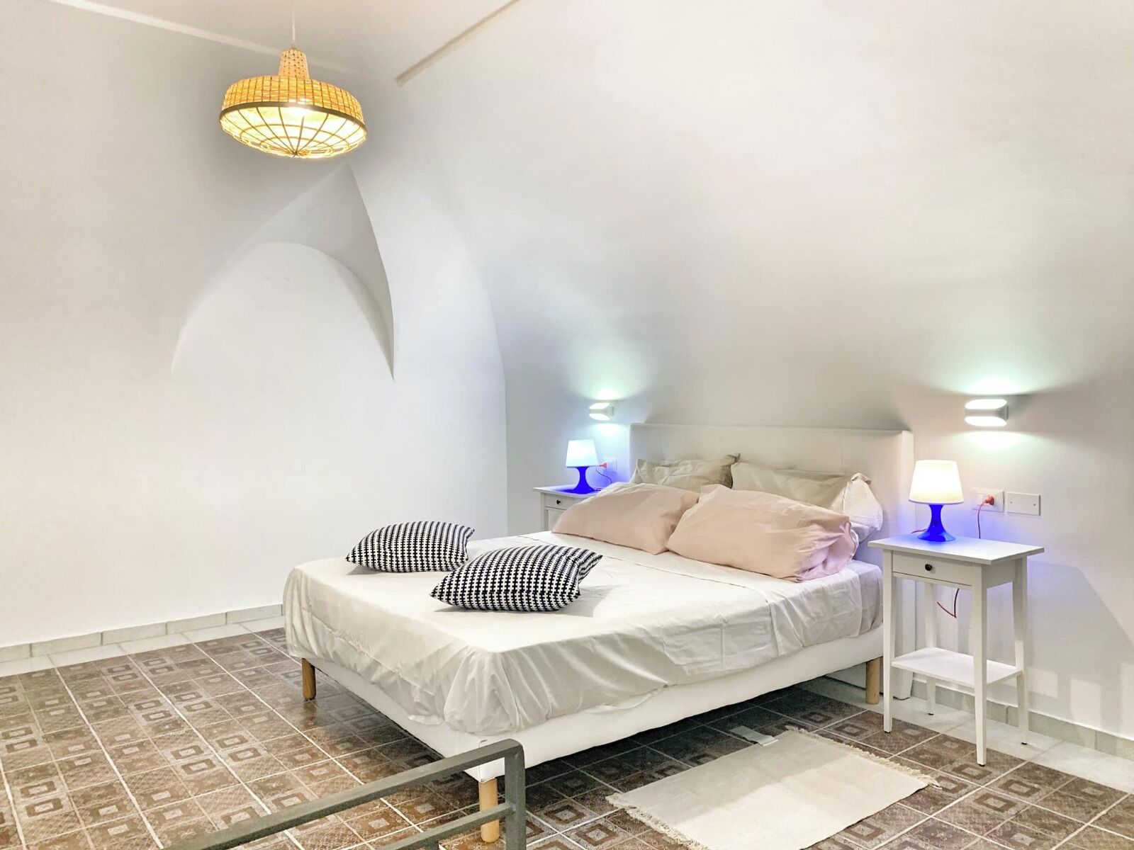 Et soveværelse med en seng, to sengeborde, en hængende lampe og gulvfliser med mønster.