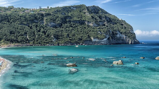 Un paysage côtier avec une plage rocheuse, des eaux turquoise et limpides, et une colline boisée.