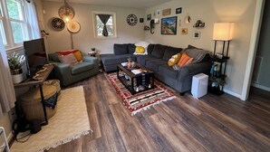 En stue med sofa, sofabord og fjernsyn. Billeder hænger på væggen, der er en lampe, og et vindue med gardiner.