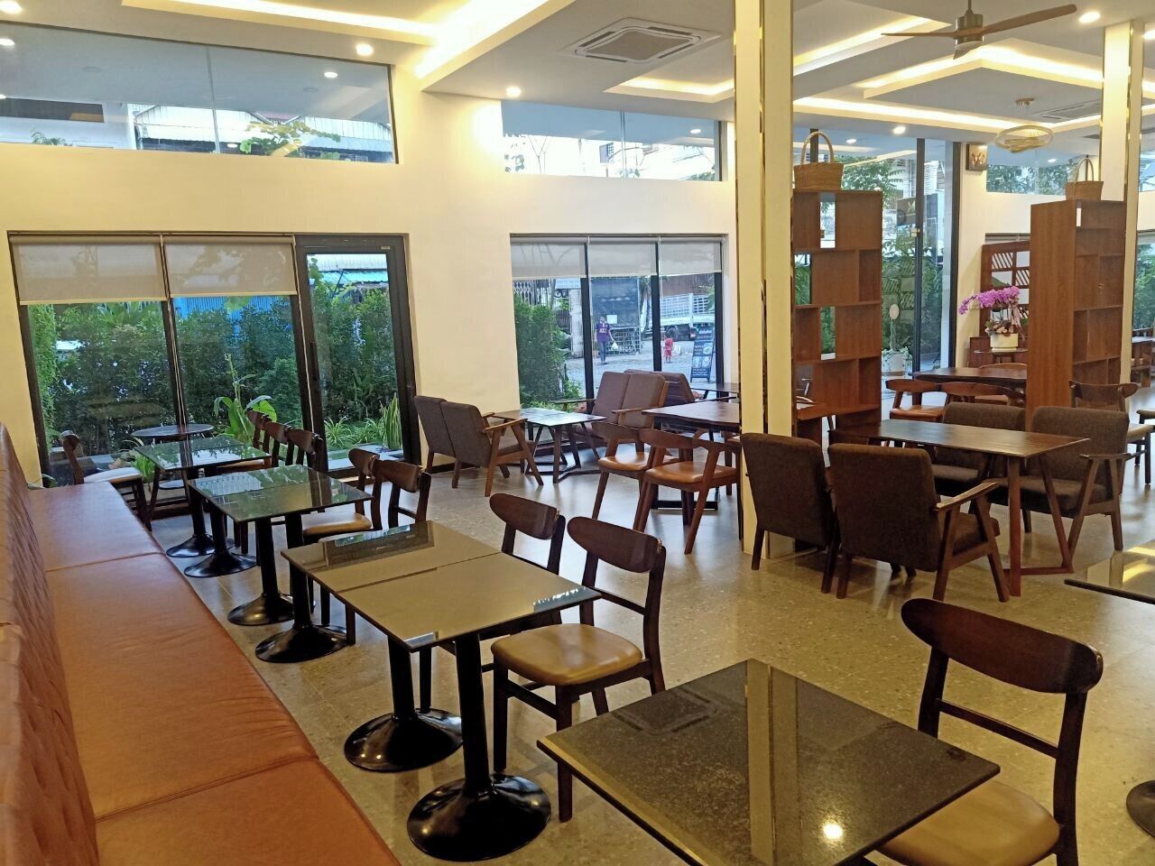 A spacious indoor area with multiple tables and chairs, large windows offering views of greenery outside, and a modern ceiling design.
